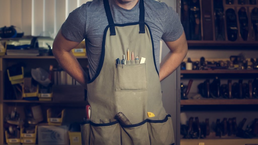 A plumber stands in front of a workbench while putting on an apron filled with tools.