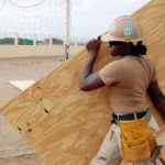 A female construction worker wearing a hard hat and work clothes, carrying a large sheet of wood across a construction site.