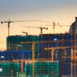 View of 10 large cranes erecting highrise buildings in an urban construction zone with the sunset in the background.